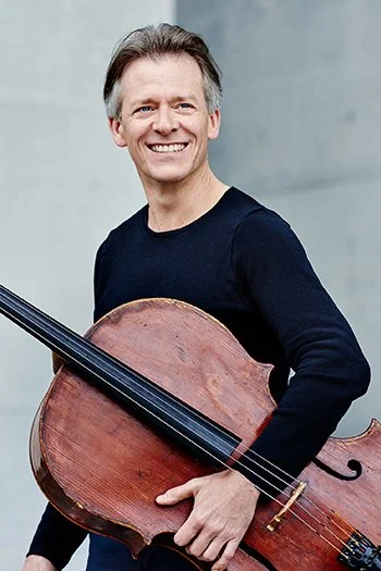 A smiling man holding a cello, standing against a plain gray background.