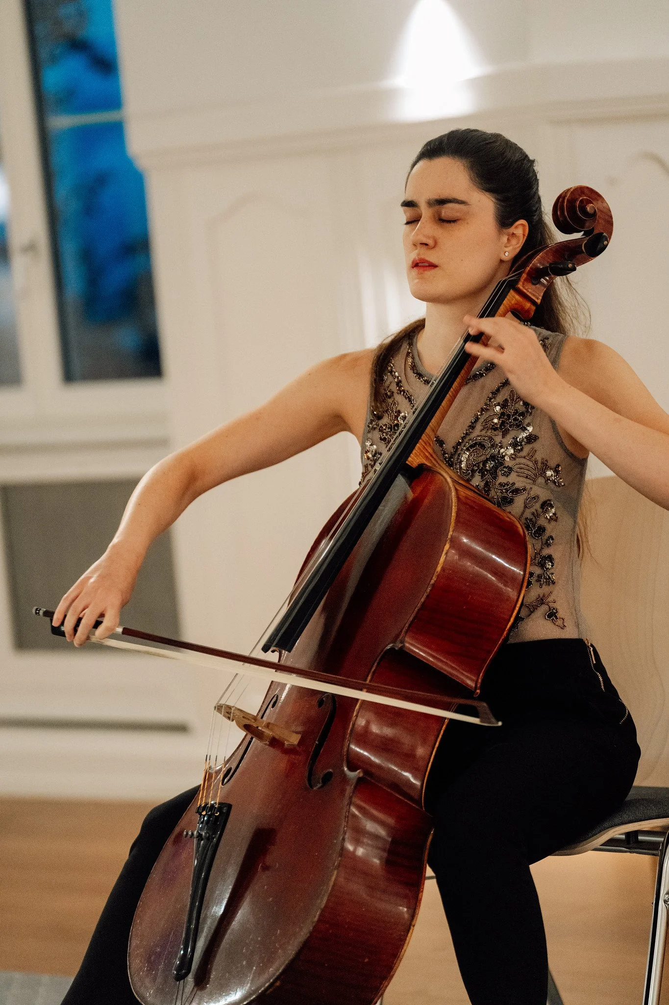 A woman with dark hair tied back, wearing a beaded, sleeveless top, and black pants, playing a brown acoustic cello in an indoor space with white walls and windows.