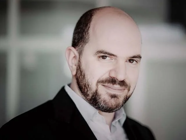 Headshot of a man with a beard, wearing a white shirt and black blazer, smiling slightly, in front of a blurred background.