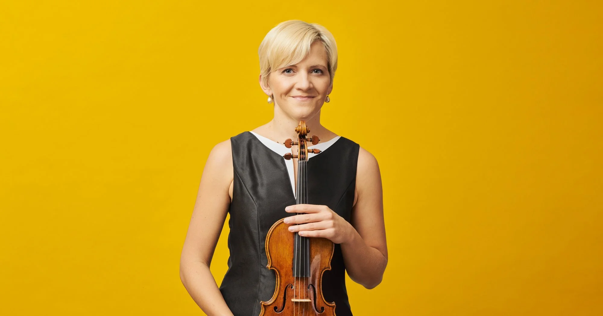 A woman with short blonde hair wearing a black sleeveless dress and pearl earrings holding a violin in front of a bright yellow background.