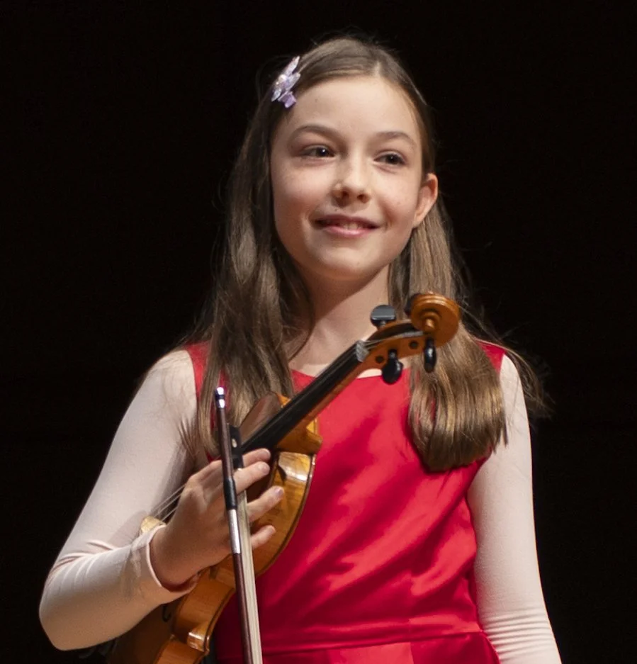 Young girl with long brown hair holding a violin and smiling, wearing a red dress and a hair clip, on a dark background.