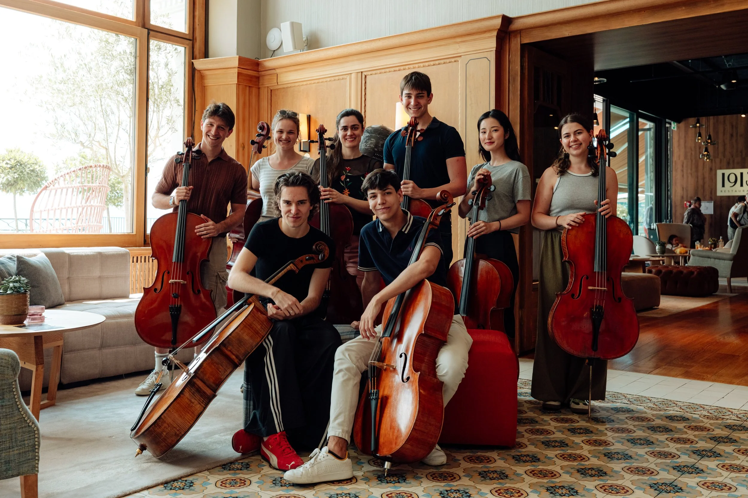 A group of nine young musicians holding cellos and standing in a cozy, well-lit indoor space with wooden interior and large windows outdoors visible.