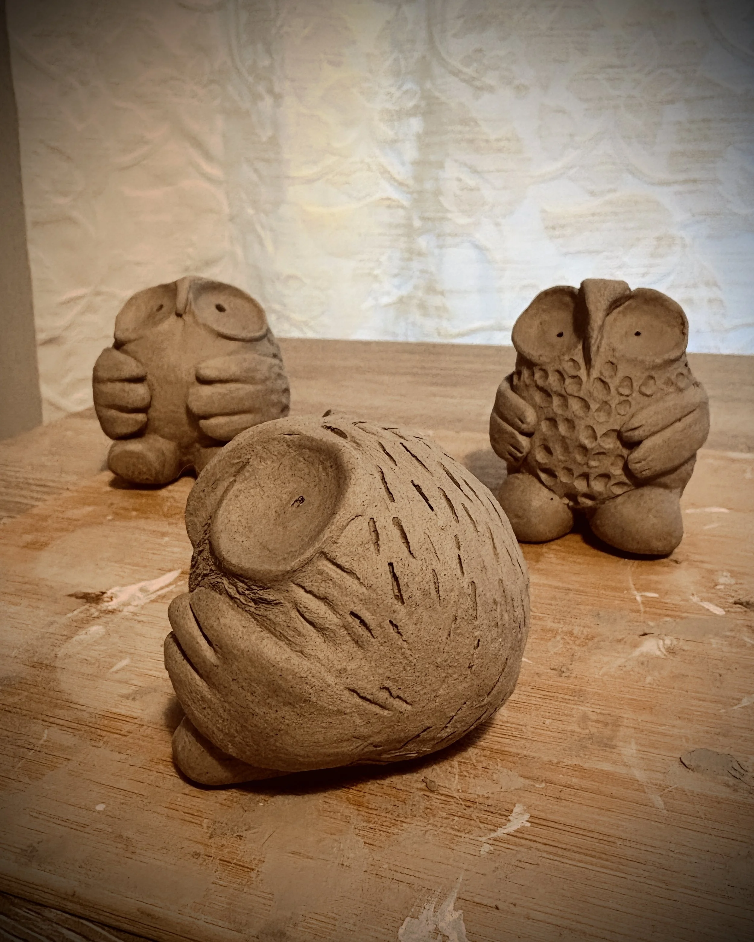 Several in progress clay sculptures of owls with big eyes and cute wings placed on a wooden surface, with a crumpled fabric backdrop.
