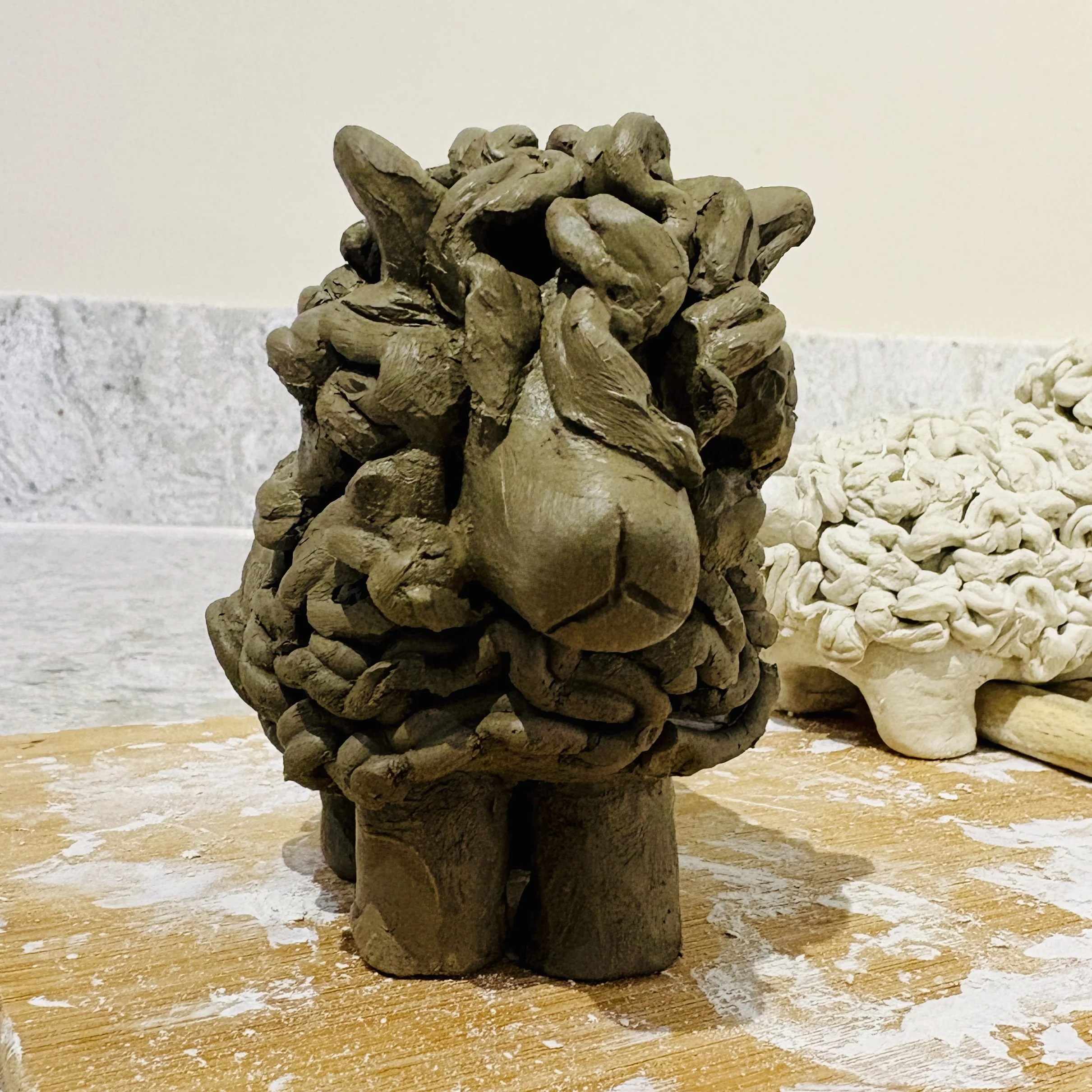 An in progress clay sculpture of a fluffy woolly sheep, on a wooden surface with scattered white powder and a light-colored wall background.