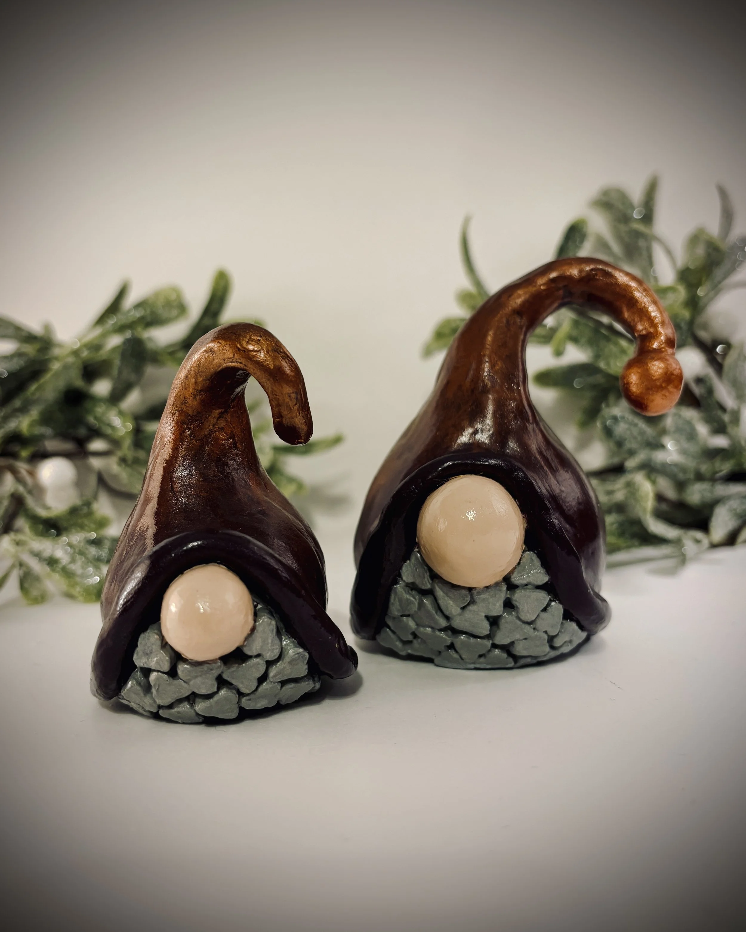 Decorative ceramic gnome figures with white beards, red hats, and grey bodies, surrounded by green foliage and white berries.