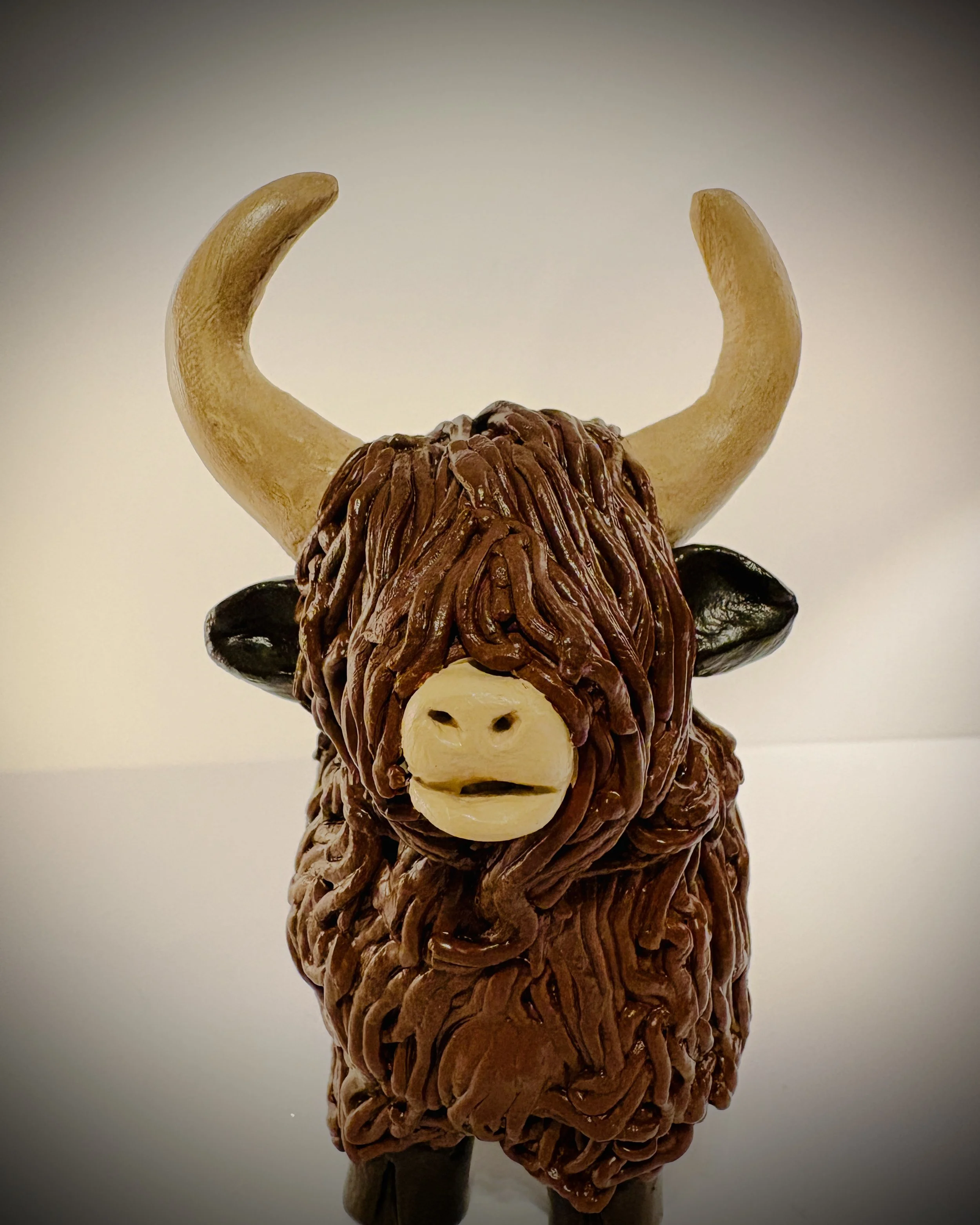 A clay sculpture of a highland cow, featuring large curved horns, black ears, a beige snout with two nostrils, and a mane made of brown material resembling hair.