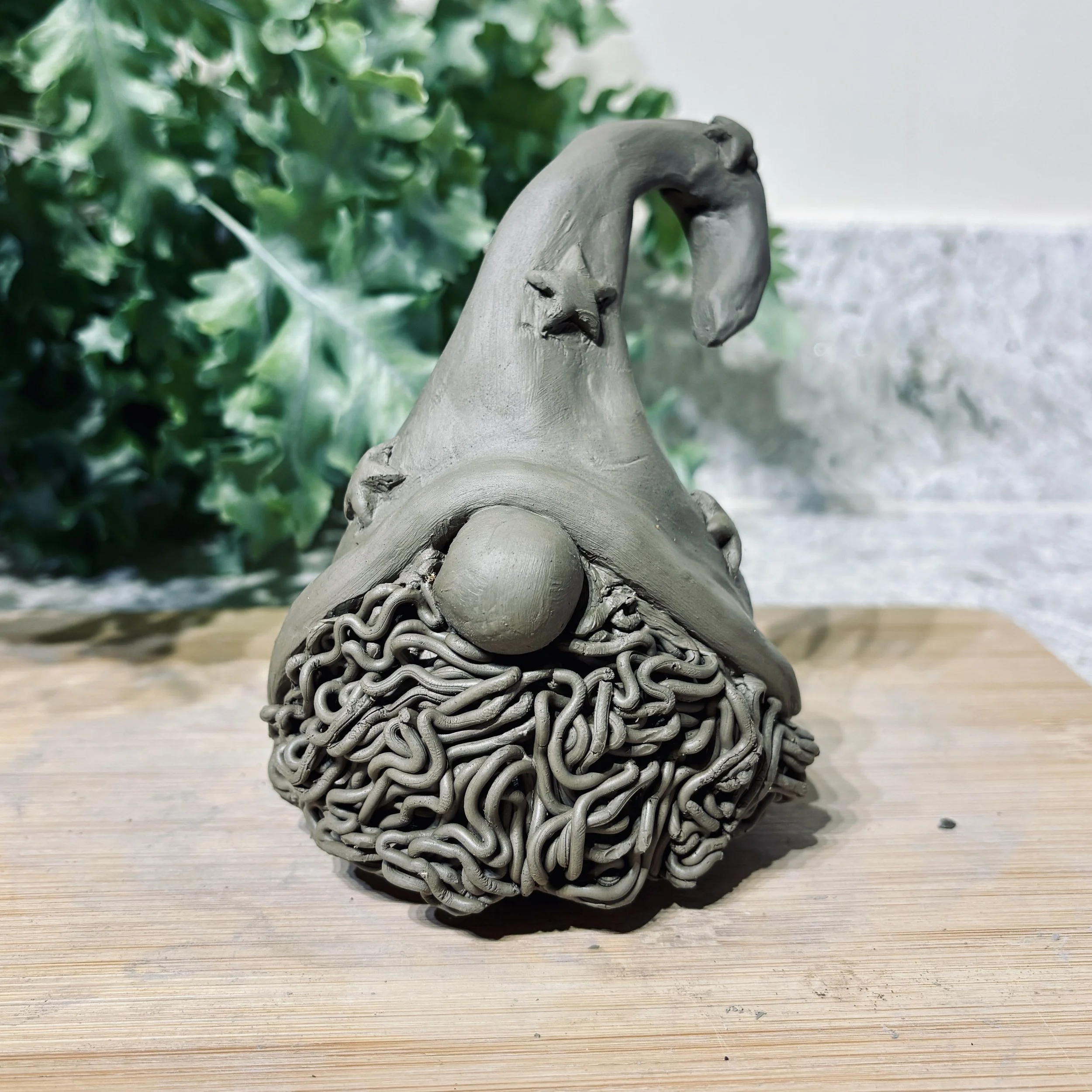 An in progess clay sculpture, of a Christmas gonk. Large hat with stars on and a big furry beard. placed on a light wooden surface with green leaves in the blurred background.