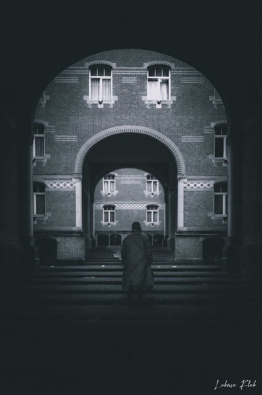 A person standing in a dark, arched passageway, looking at a multi-story brick building with several windows.