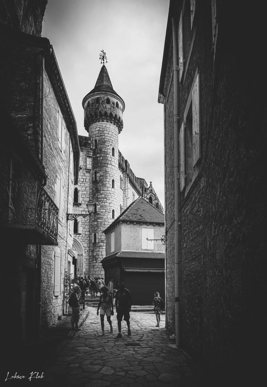 A black and white photo of a narrow street leading to a castle with a tall, rounded tower topped with a weather vane. People are walking along the cobblestone street.
