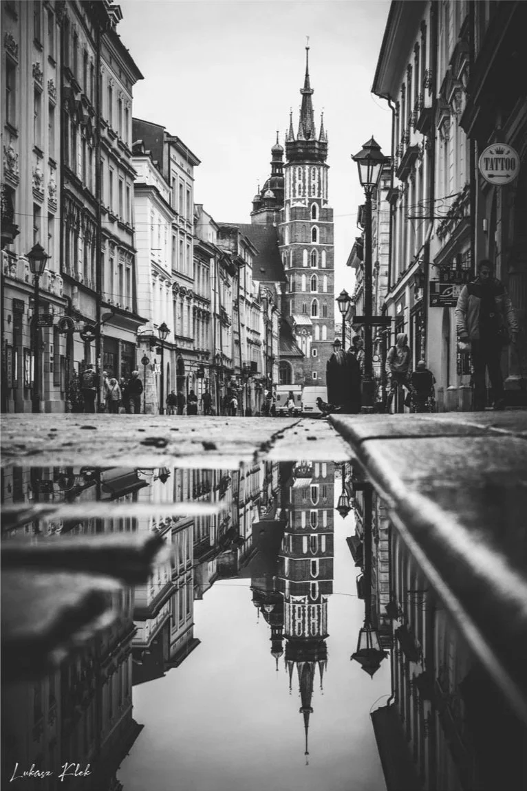 Black and white photo of a European city street with historic buildings and a tall tower reflecting in a puddle on the sidewalk.