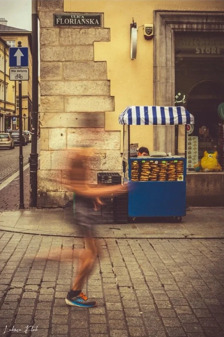 A blurred person walking on a cobblestone street next to a blue and white striped street vendor cart selling food, with a yellow building and shop window in the background on Florian Street.