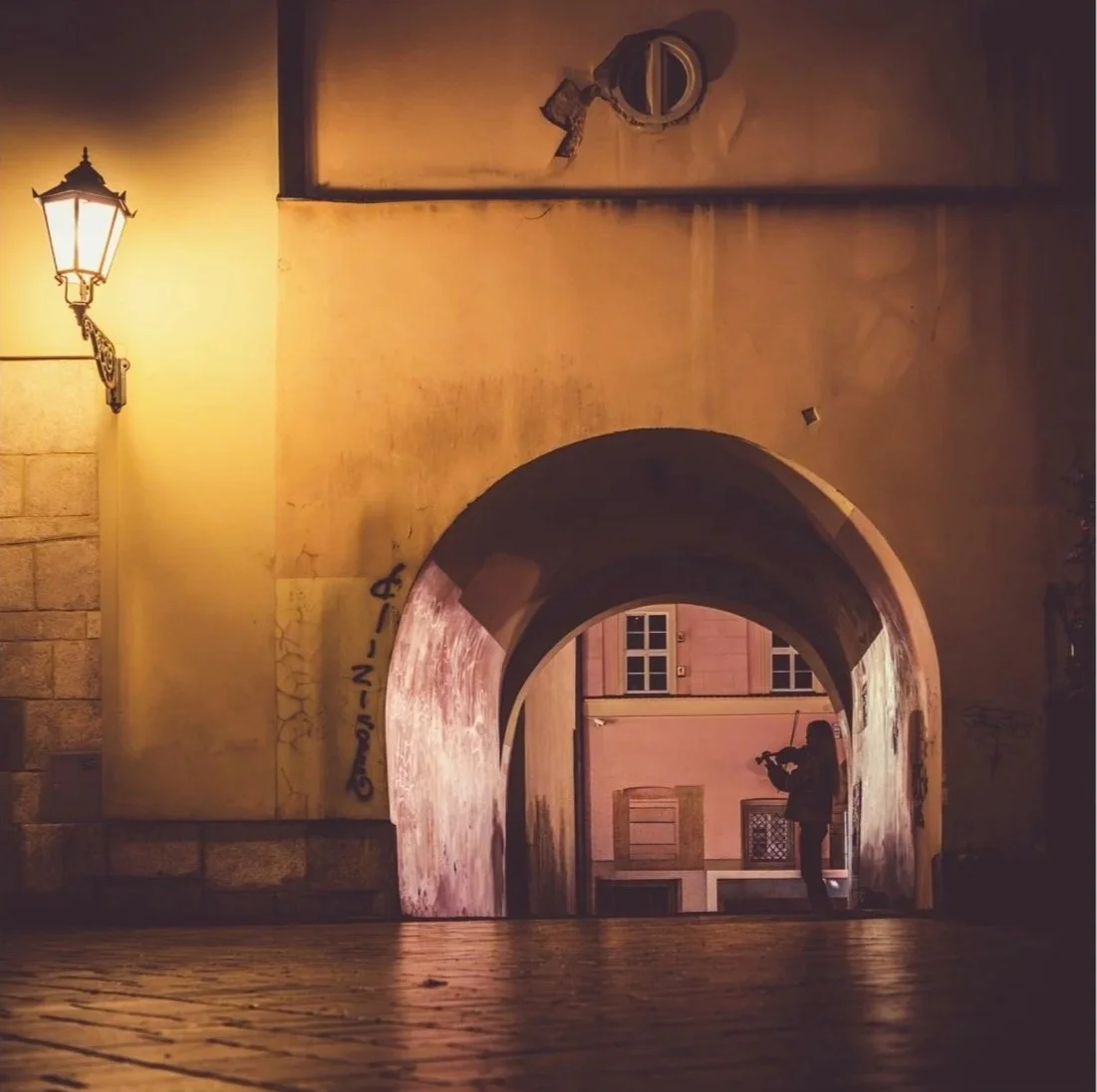 Night scene of a person taking a photograph under a lit archway in a European-style street, with warm yellow lighting and old buildings.