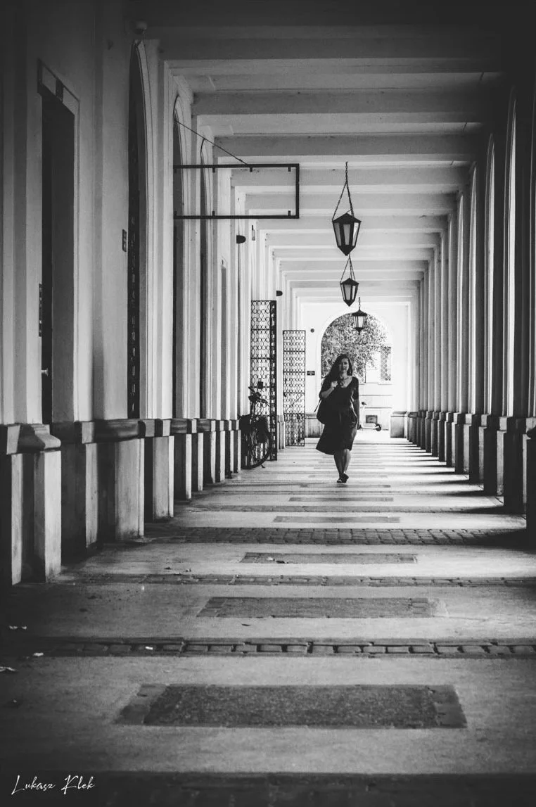 A black and white photo of a woman walking through an arched walkway with columns on both sides. The ceiling has hanging lantern-style lights, and there are decorative panels and a bicycle near the wall. The pathway has a patterned surface, and there