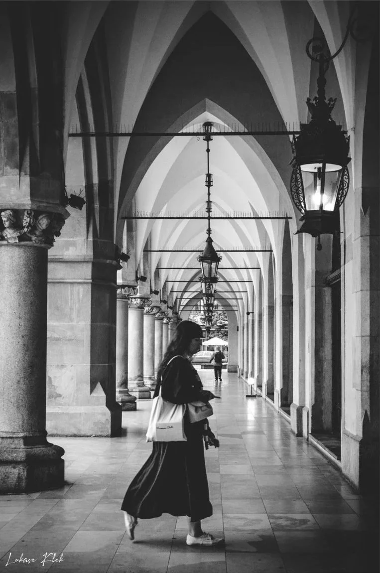 A woman walking in a covered outdoor walkway with arched ceilings, ornate columns, and hanging lanterns, in black and white.