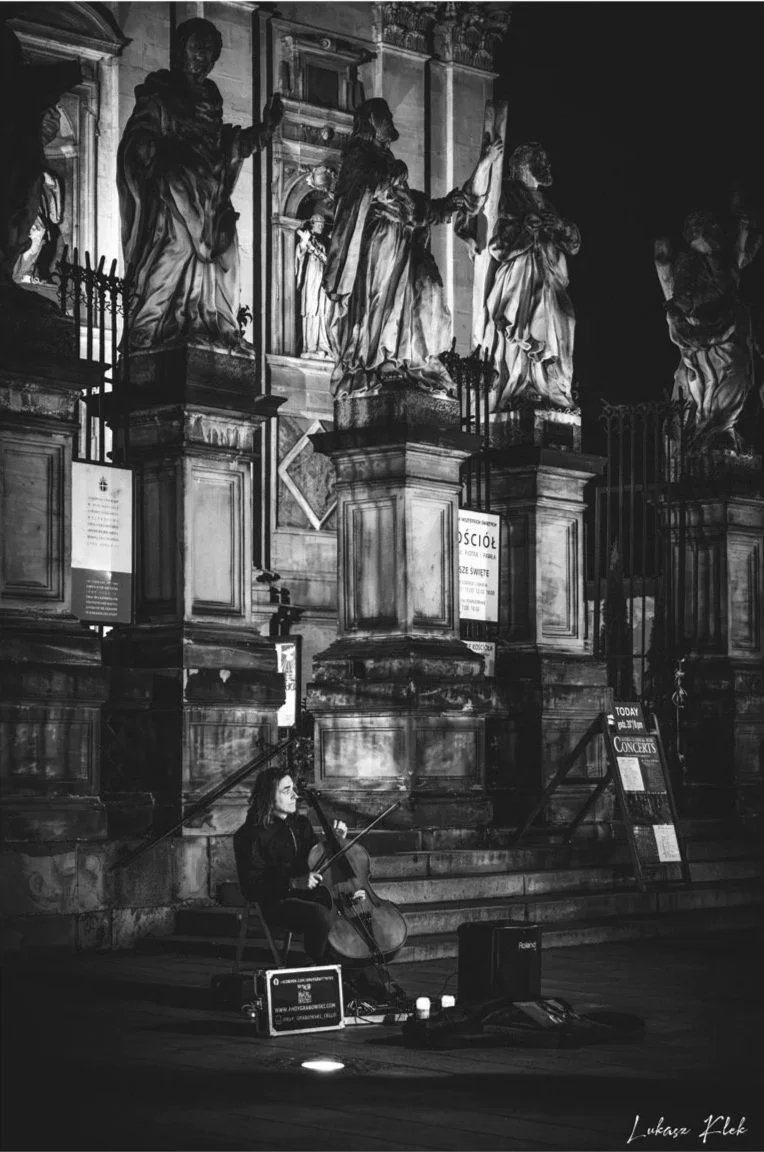 A woman playing a cello and singing in front of an ornate, historical building with statues and columns, illuminated at night.