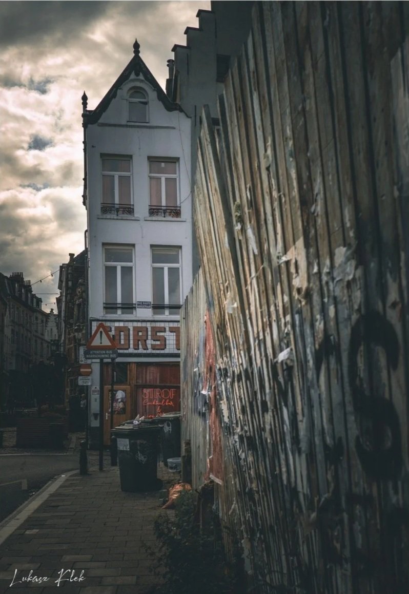 A narrow street scene in a city with an old building with ornate roofline, graffiti-covered metal fence on the right, business sign on a storefront, and a cloudy sky overhead.