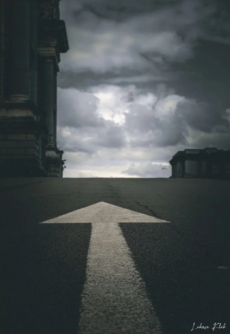 An upward pointing white arrow painted on a dark asphalt street, with cloudy sky overhead and buildings on either side.