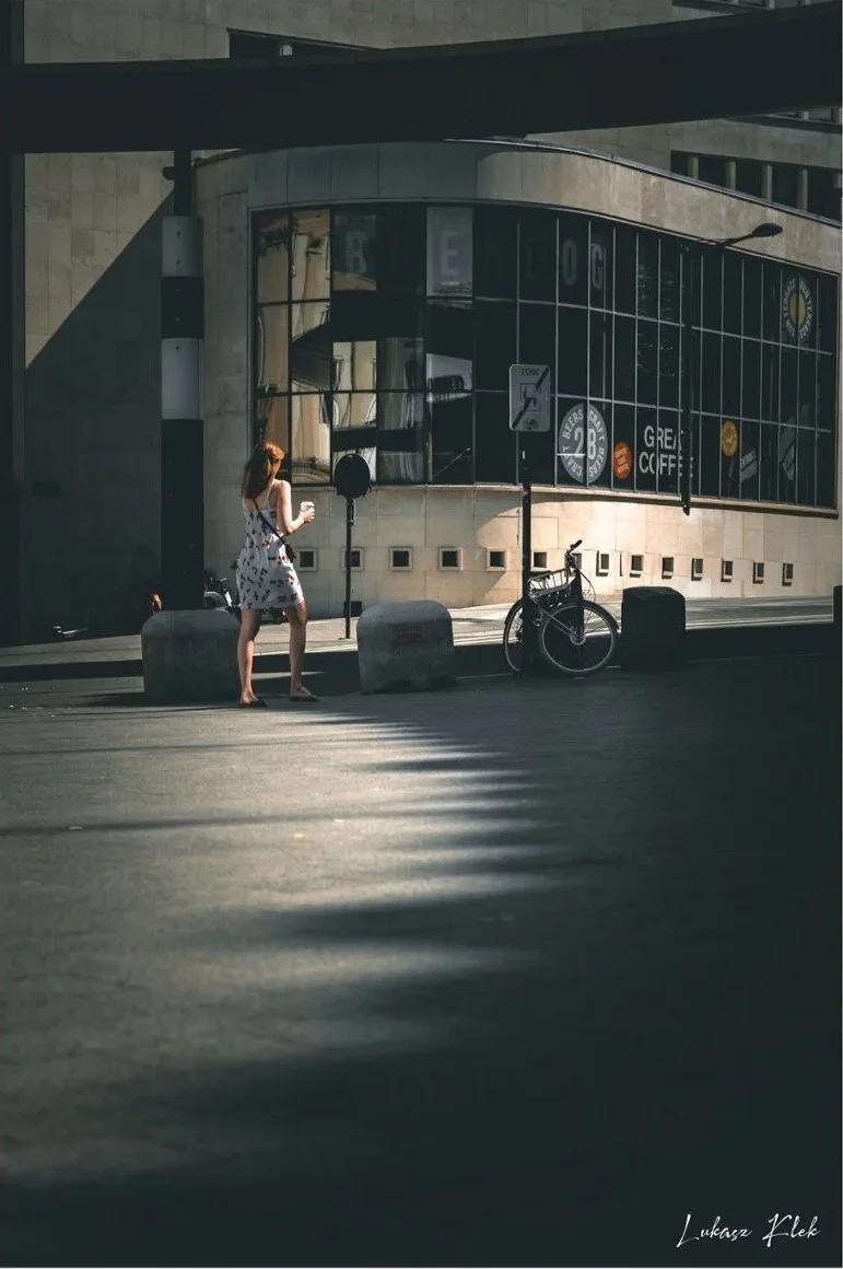 Woman with red hair in a white dress with black and red patterns standing on a city sidewalk, holding a coffee cup; bicycle parked nearby; modern building with reflective glass windows in the background.