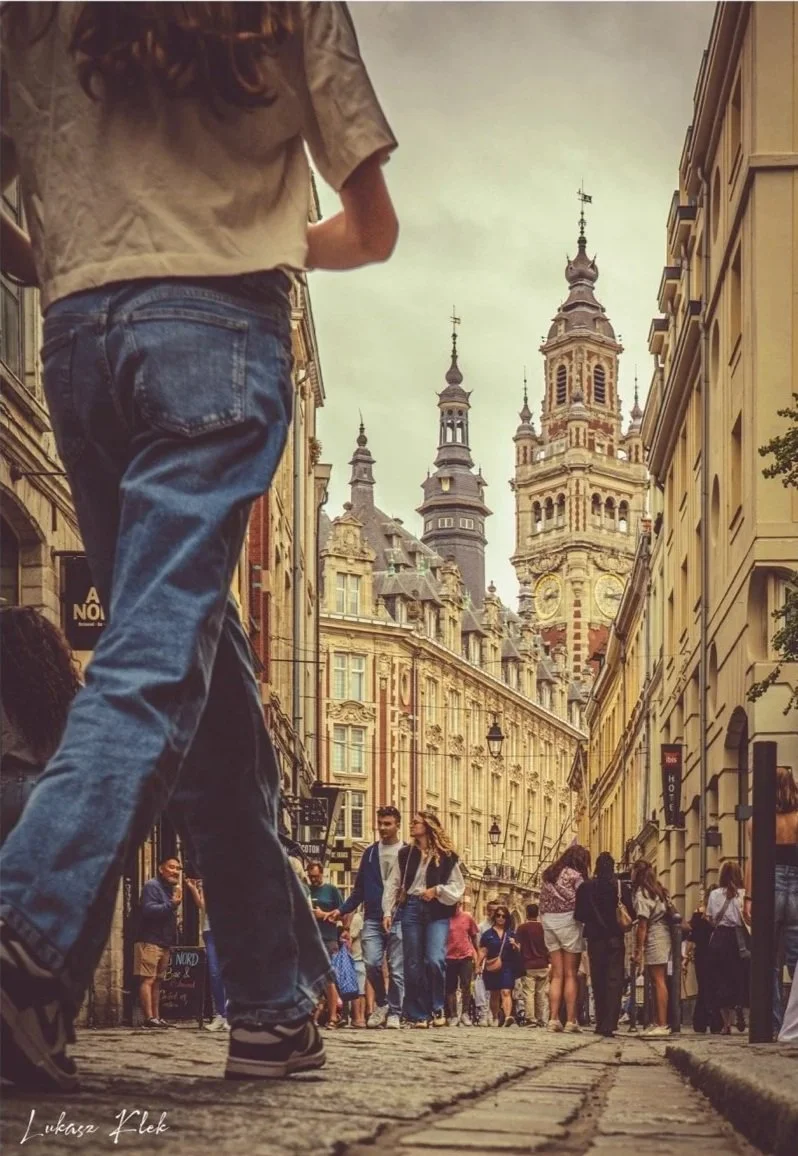 A bustling city street scene with historic architecture, people walking, and a person in jeans and sneakers in the foreground.