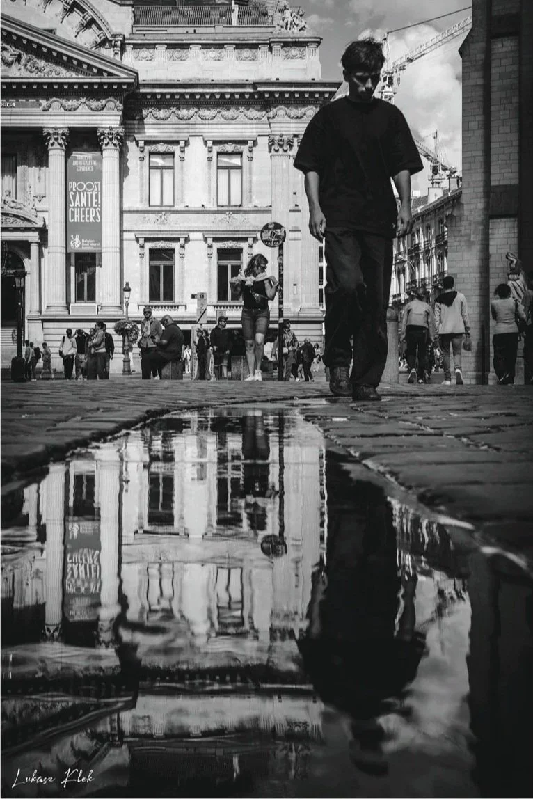 A black and white photo of a city street with a historic building in the background. A large puddle on the cobblestone street reflects the building and people walking by. A young man in dark clothing walks toward the camera, with other pedestrians, i