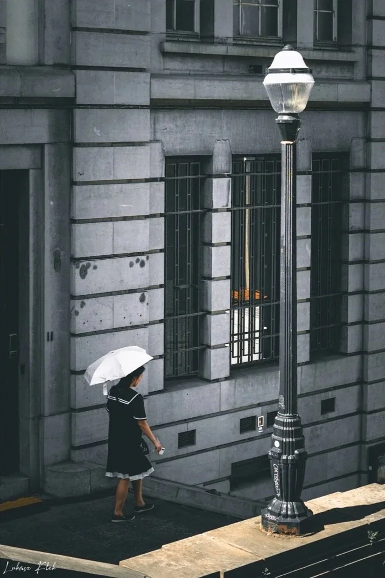 A woman in a black dress holding a white umbrella walking on a city sidewalk near a stone building with barred windows and a streetlamp.