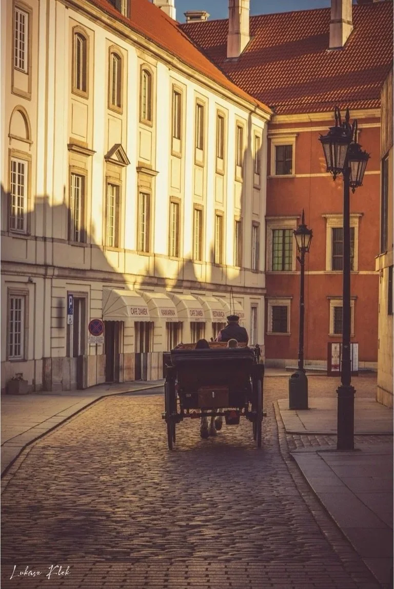 A horse-drawn carriage traveling down a cobblestone street lined with European-style buildings and street lamps during sunset.