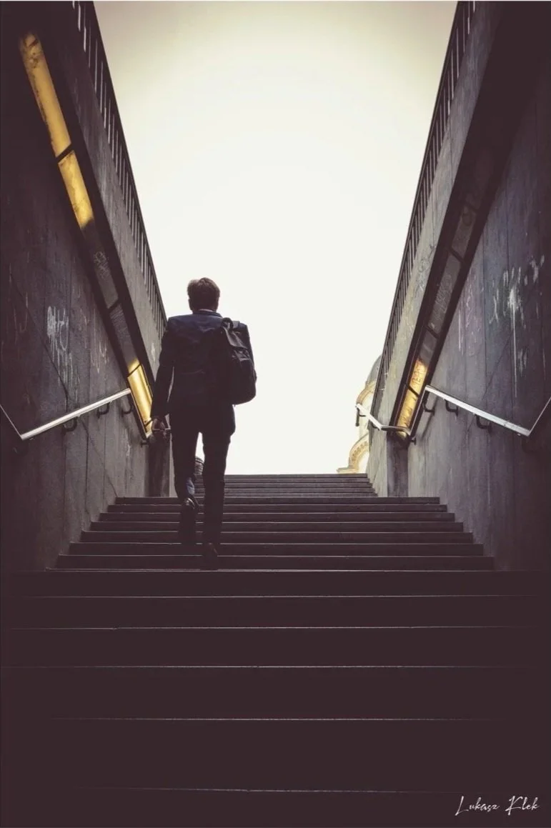 A person in a dark suit with a backpack walking up stairs towards a bright light at the top of an underground passage or subway entrance.