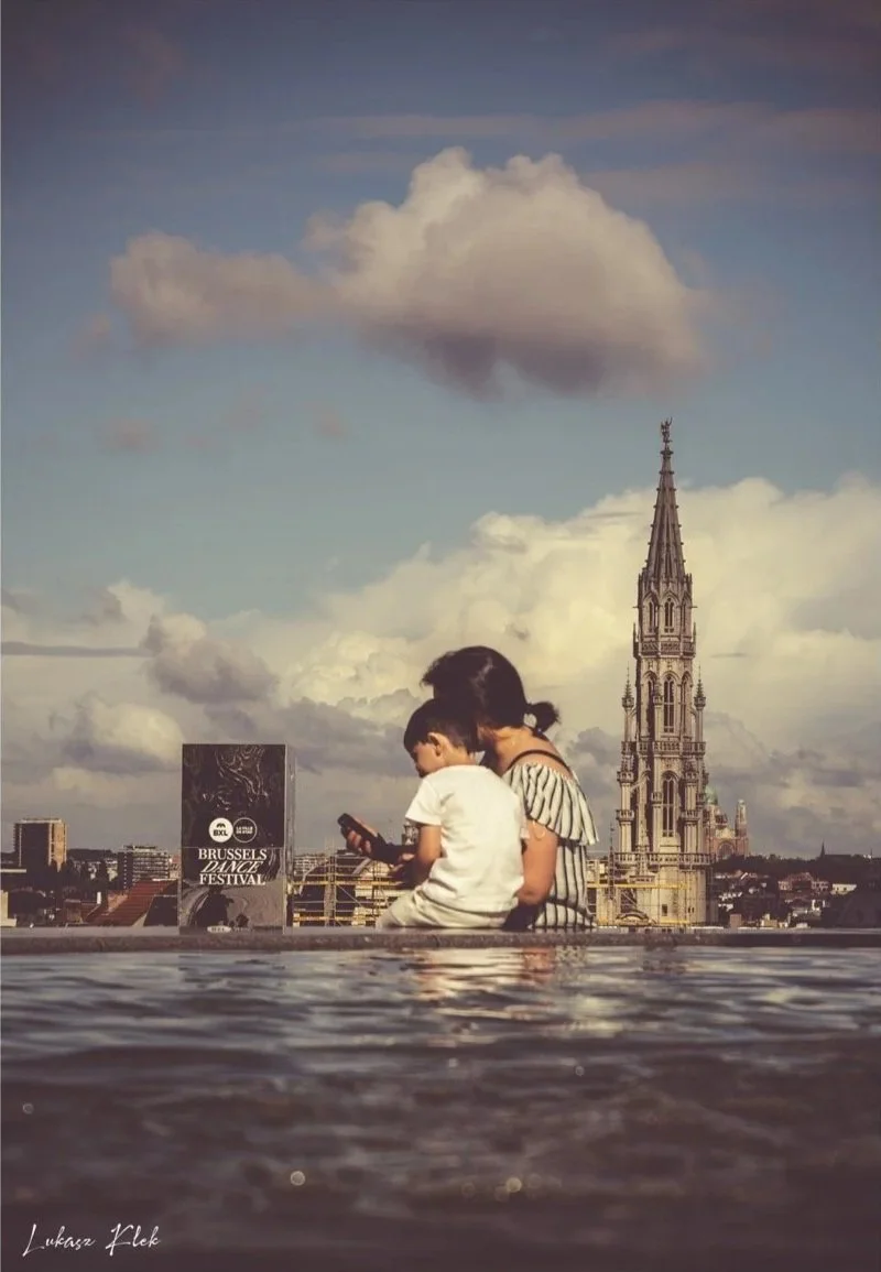 A woman and two children sitting near a water body, looking at a phone, with a cityscape and a tall church spire in the background during daytime.