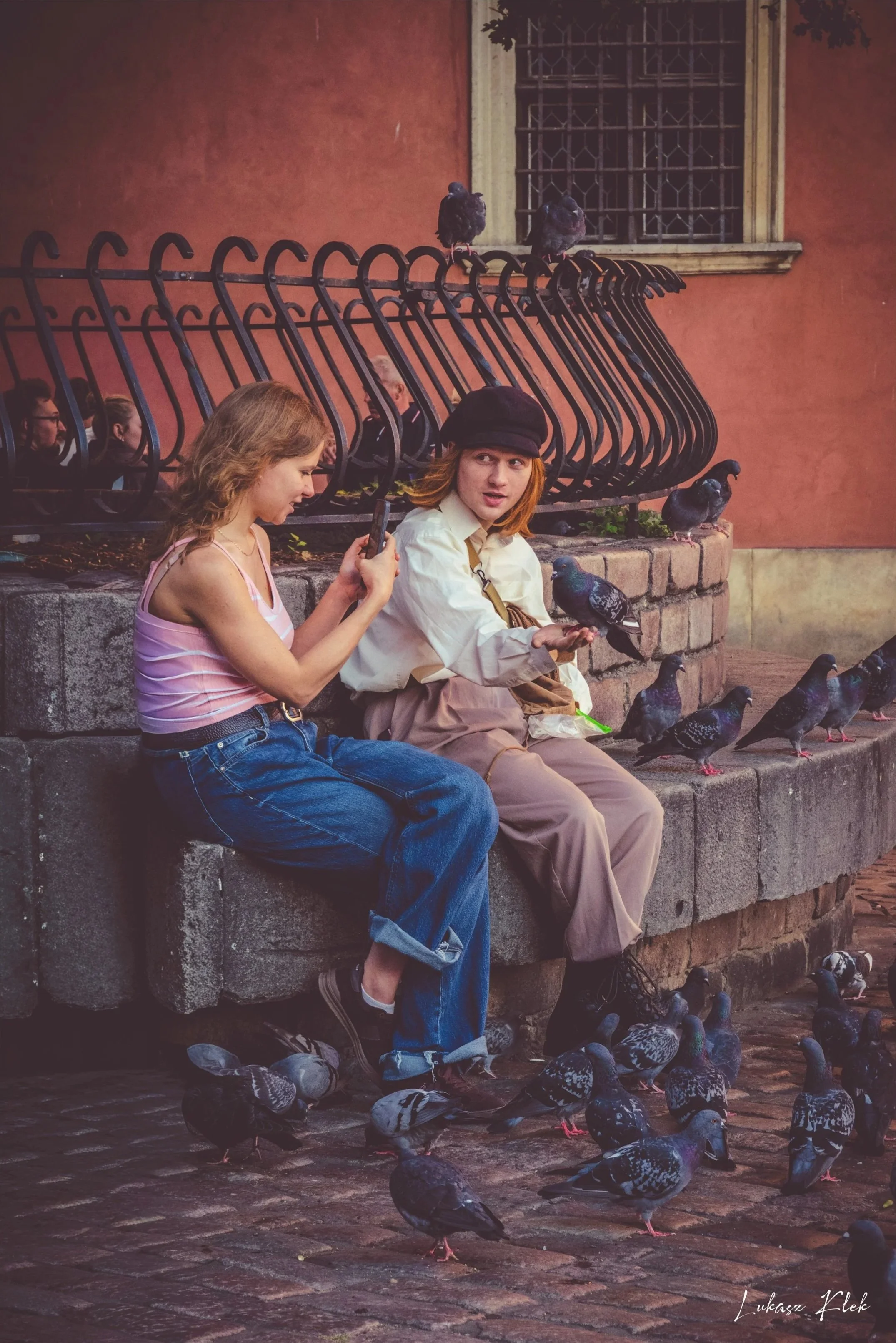 A young woman and a young man sitting on a stone ledge, surrounded by pigeons. The woman has long curly hair, wearing a pink striped tank top and jeans, looking at her phone. The man has shoulder-length hair, wears a black beret and beige clothing, h