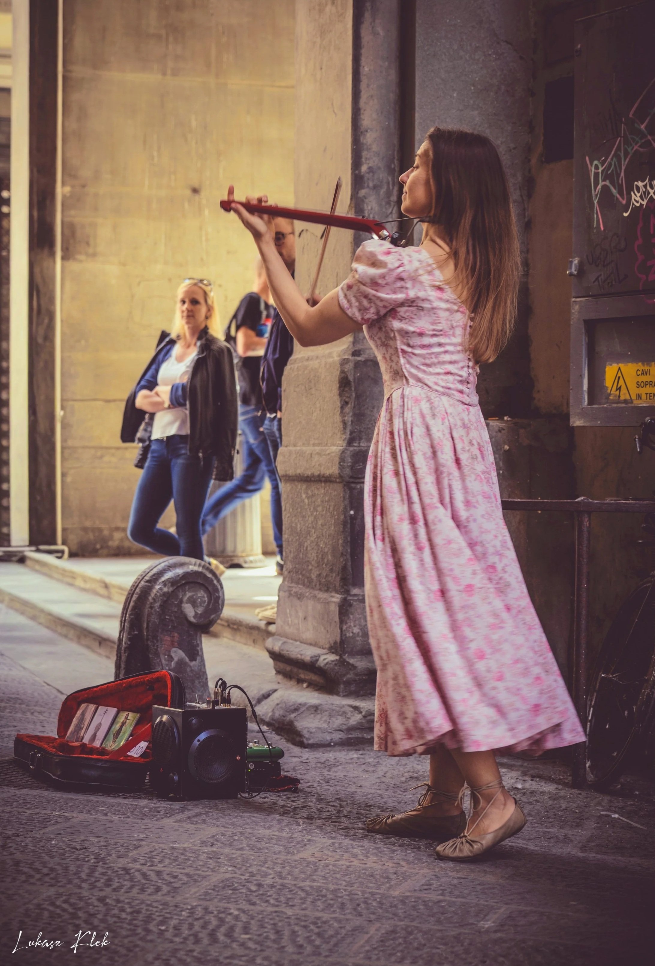 A woman in a pink floral dress is playing a violin on a city street at night. She is standing in front of an old stone building, with a small portable speaker and a box of money on the ground nearby. Two women and a man are watching her, standing aga