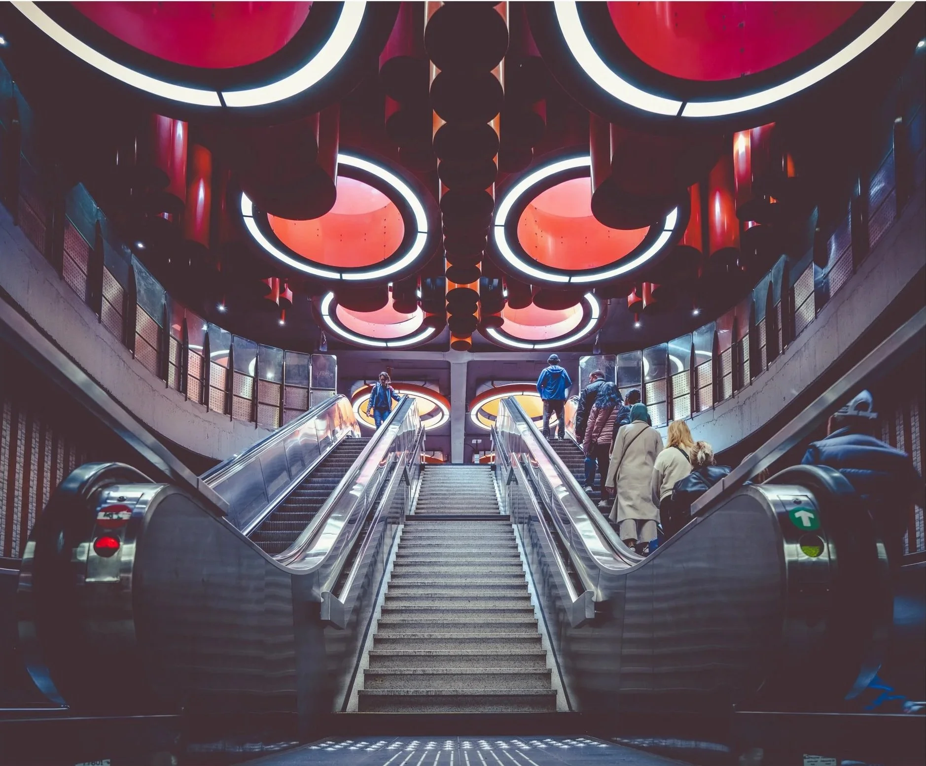 People walking on escalators in a modern shopping mall or transit station with a colorful ceiling featuring large circular lights and red decorative elements.