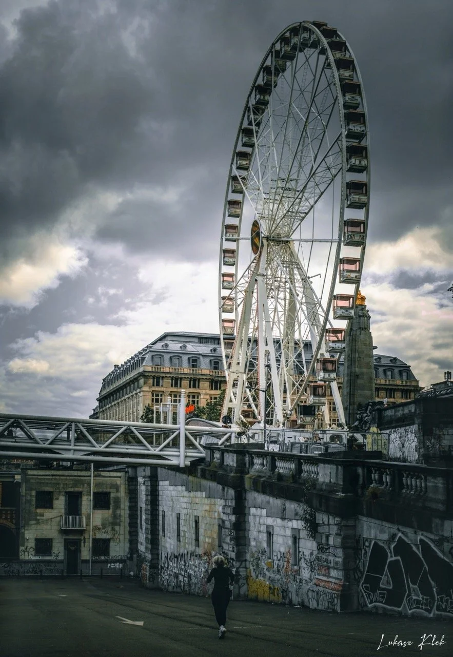 A large Ferris wheel overlooking a city with classical architecture, under dark cloudy skies, with a lone person jogging on a street below covered in graffiti.