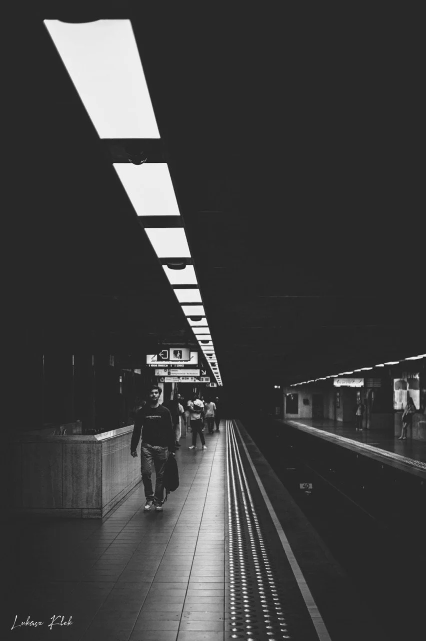 Black and white photo of a subway station platform with a few people walking. The platform has overhead fluorescent lights and safety lines near the edge. A sign with arrows and symbols hangs above in the distance.