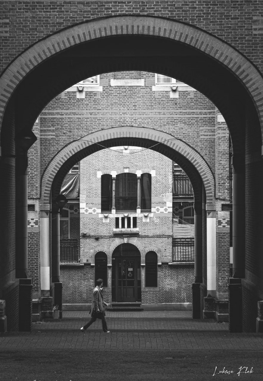 A person walking in an alleyway framed by brick arches, with a brick building with windows in the background.