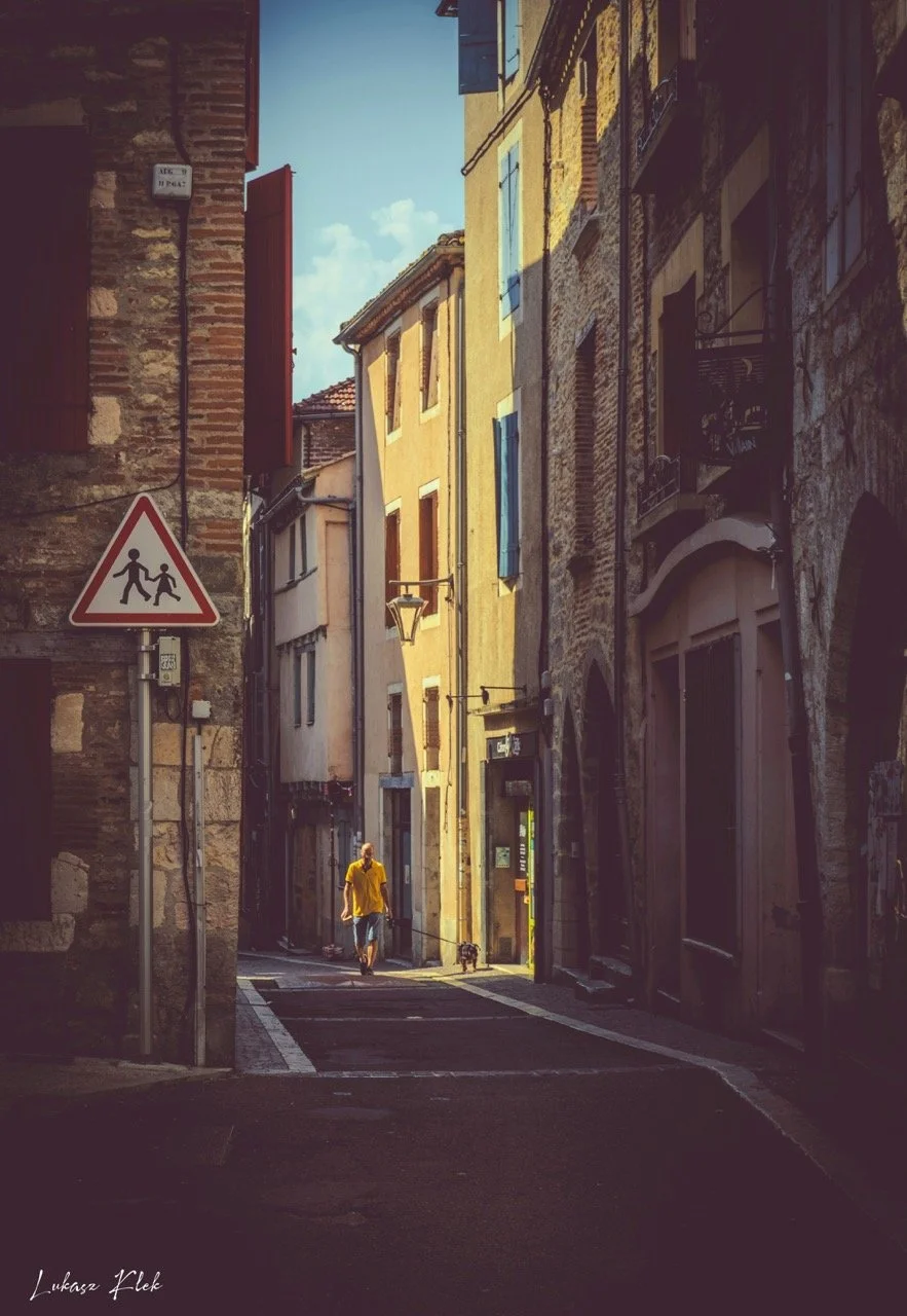 A narrow European street with old brick and stucco buildings, a person in a yellow shirt walking a dog, a pedestrian crossing sign, and decorative street lamps