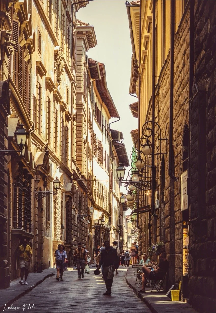 A narrow European street bustling with pedestrians, lined with historic stone and brick buildings. Some people sit at outdoor tables, and vintage-style street lamps hang above.