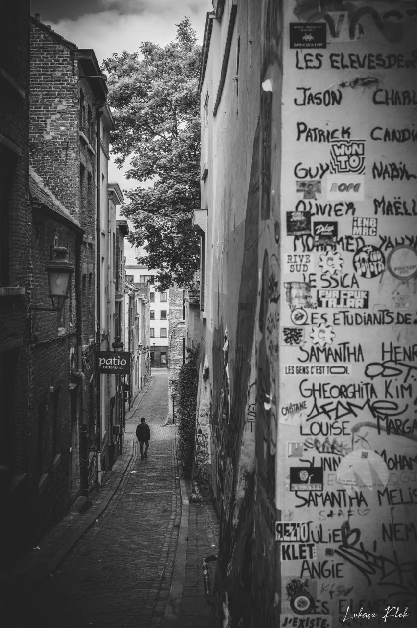A black and white photo of a narrow alleyway with a person walking away from the camera. The alley is lined with buildings on both sides, with graffiti and stickers on the right wall. A sign for a pottery shop is visible on the left.