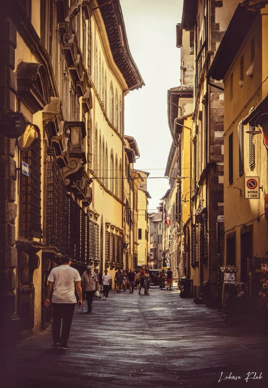 A narrow European street lined with tall, historic buildings with ornate architectural details, pedestrians walking and shopping, and warm, muted lighting.