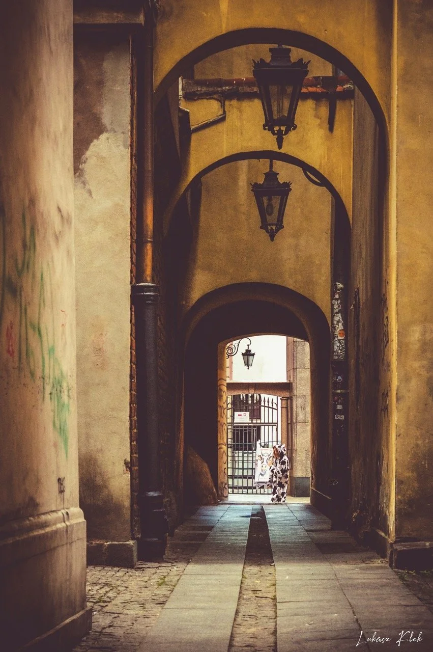 Narrow alleyway with yellow walls, arched doorways, and hanging lanterns, leading to a gate with a person in a cow-patterned outfit near the back.