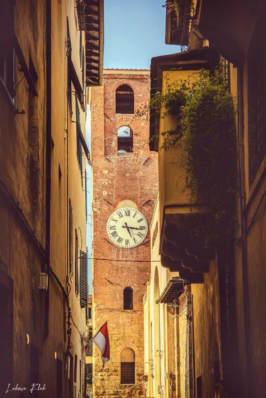 A narrow street in Italy with tall, yellow and brown buildings on both sides, a flag hanging from a pole, and a brick clock tower with a large clock showing 2:13, sunlight casting shadows.
