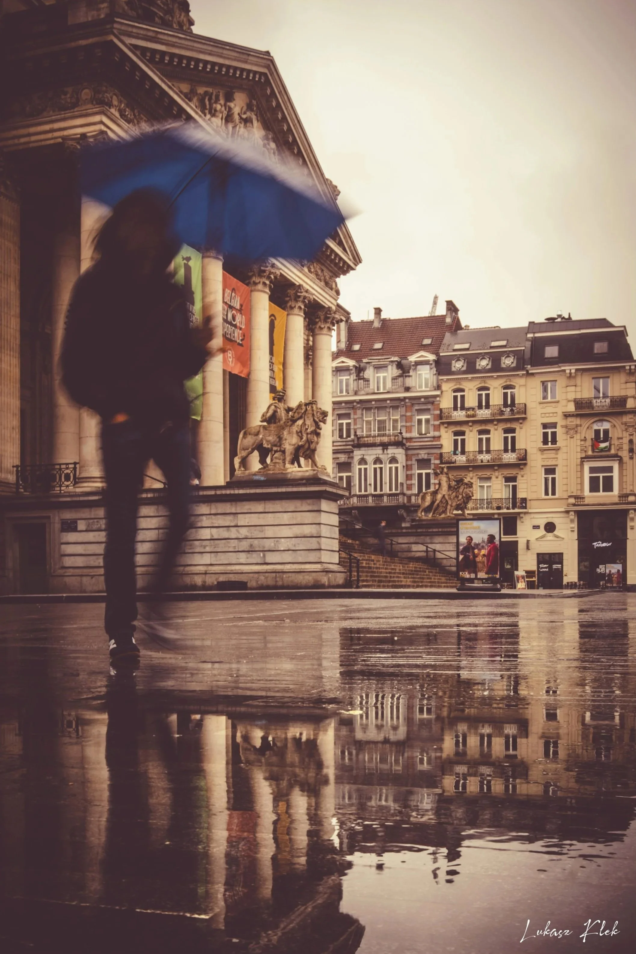 A person walking with an umbrella in front of a historic building with classical architecture and sculptures, reflected on a wet ground.