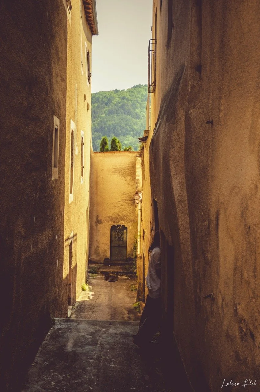 A narrow alleyway between tall, old yellow buildings with small windows, leading to a small courtyard with a fountain and surrounded by greenery, overlooking distant green hills.