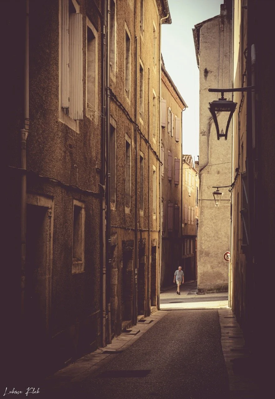 A narrow alleyway between tall, old buildings with weathered walls in a European city, illuminated by sunlight with a person walking in the distance.
