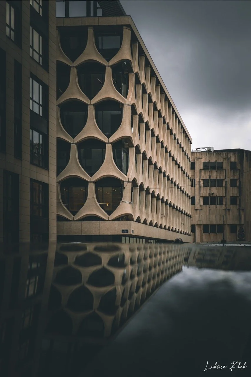 A modern multi-story building with a distinctive honeycomb-like facade next to a reflective water surface, under dark cloudy skies.