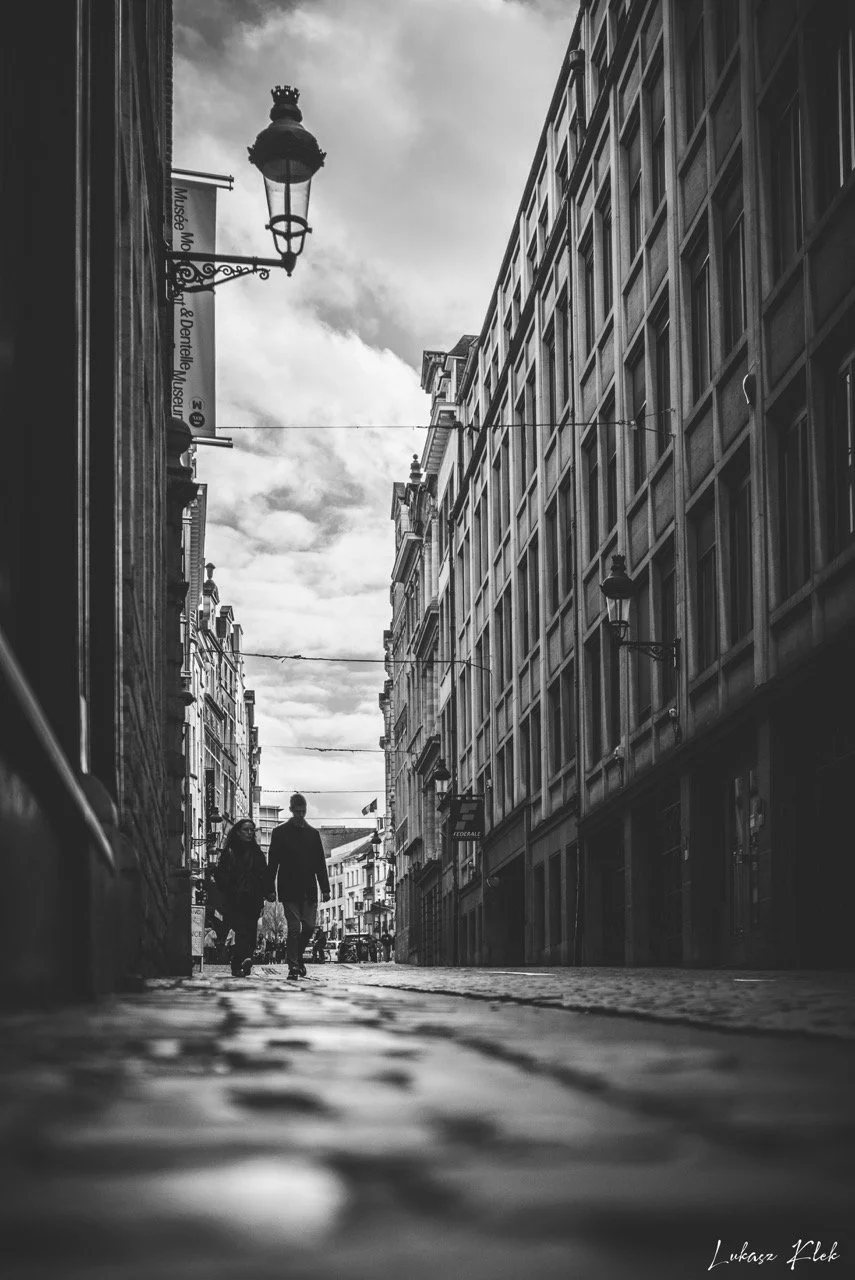 Black and white photo of a city street with two people walking on cobblestone pavement, tall buildings lining the street, vintage street lamps, and cloudy sky overhead.