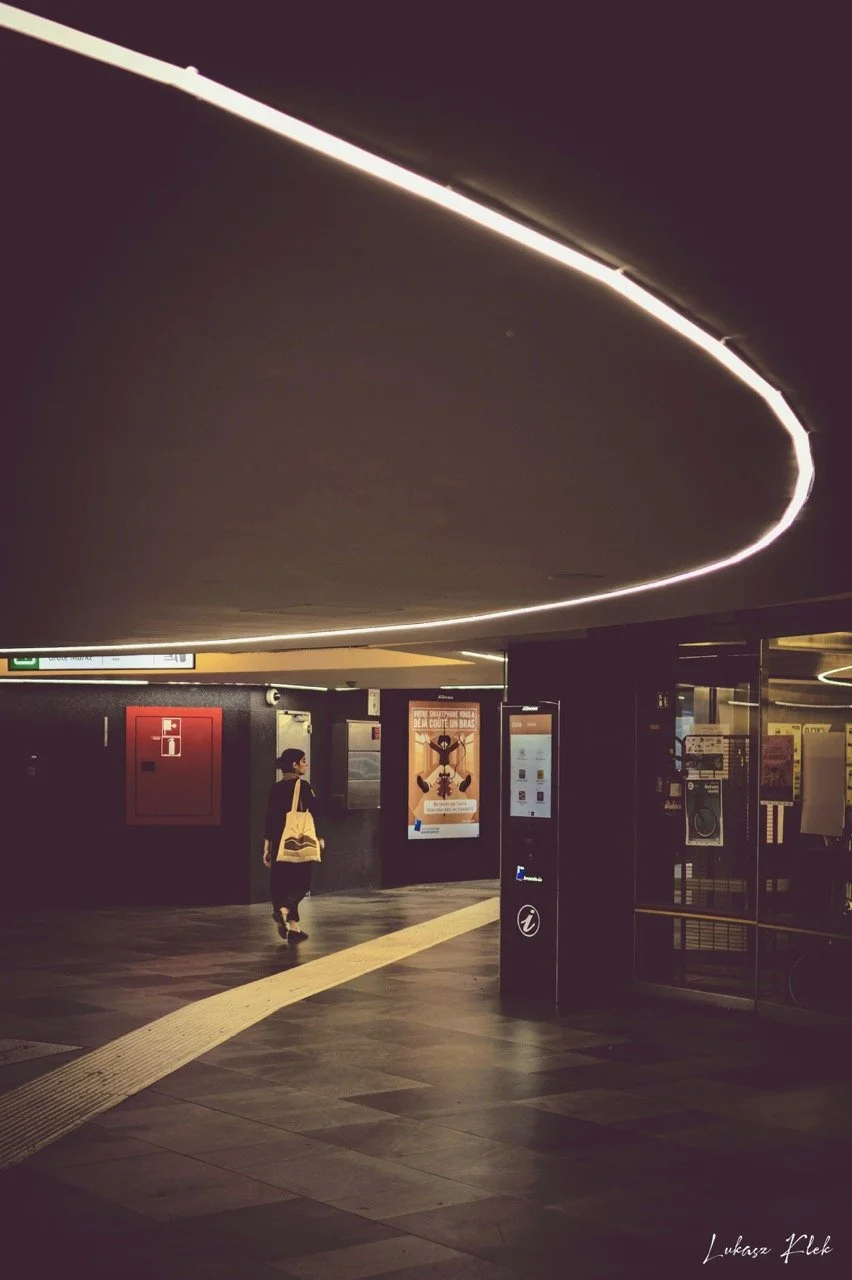An underground subway station with a person walking past illuminated advertising posters and a large circular ceiling light.