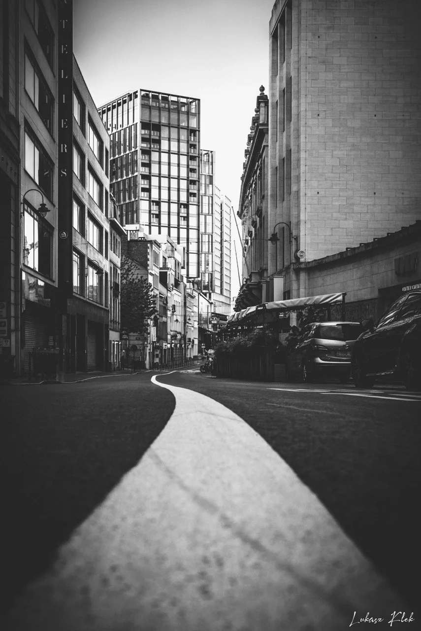 Black and white photo of a city street with tall buildings on both sides. A curved white line runs down the center of the road, with parked cars along the curb. The skyline features modern glass and concrete buildings.