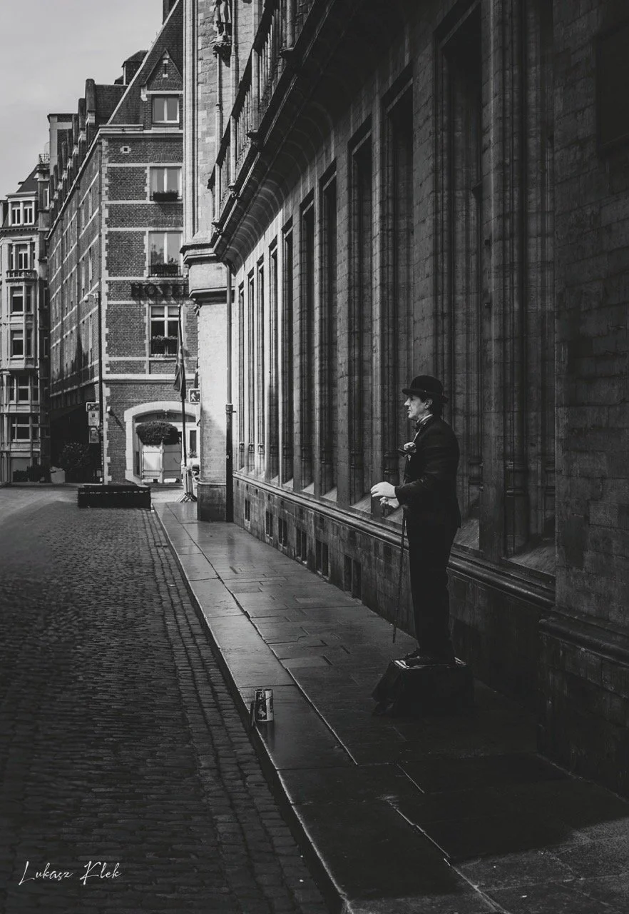 A street scene in a European city with old buildings. A man dressed in vintage clothing, including a bowler hat, is standing on a small box on the sidewalk, appearing to perform or speak with a harmonica. The street is lined with cobblestones and wet