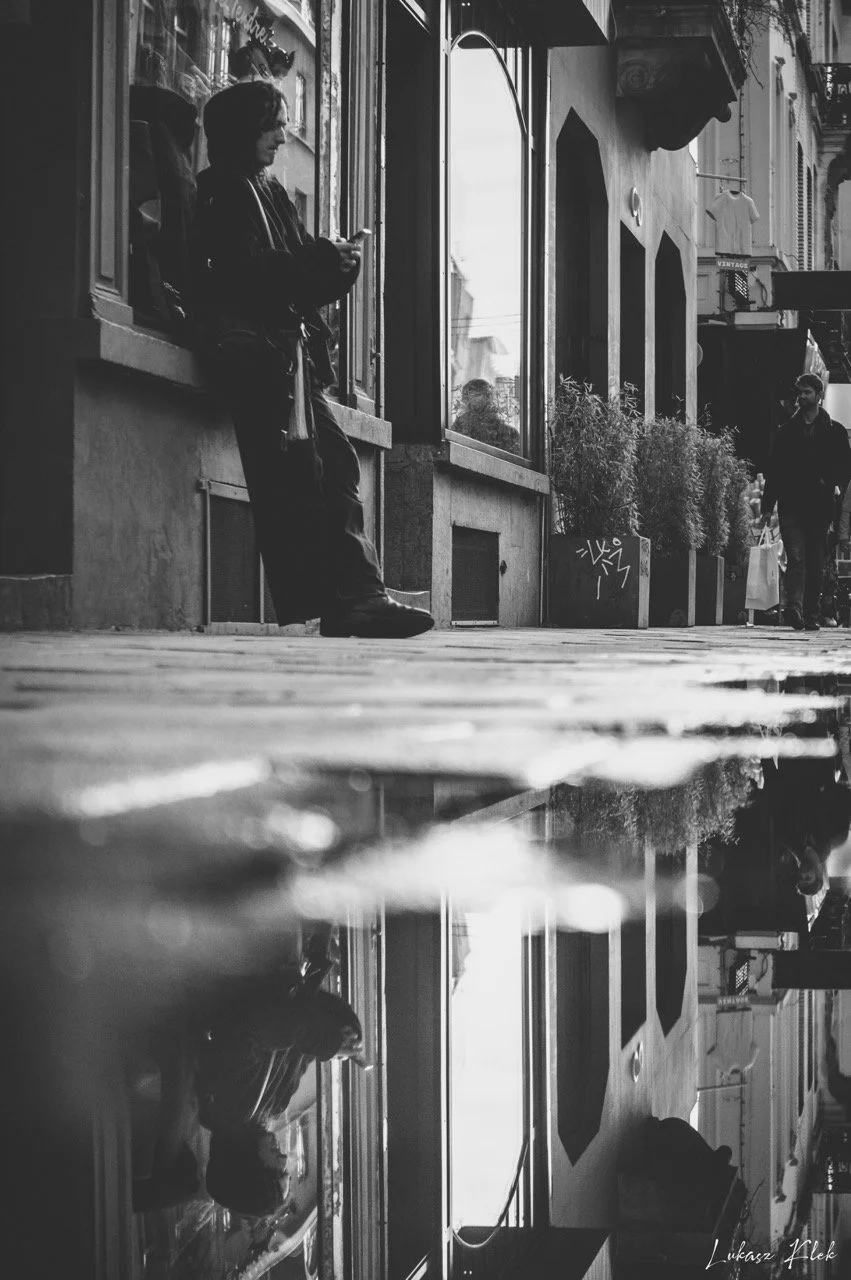 A man leaning against a window sill on a city sidewalk, looking at his phone. The street has buildings with storefronts and other pedestrians walking by. The photo is in black and white, with a reflection of the scene seen on the wet pavement.