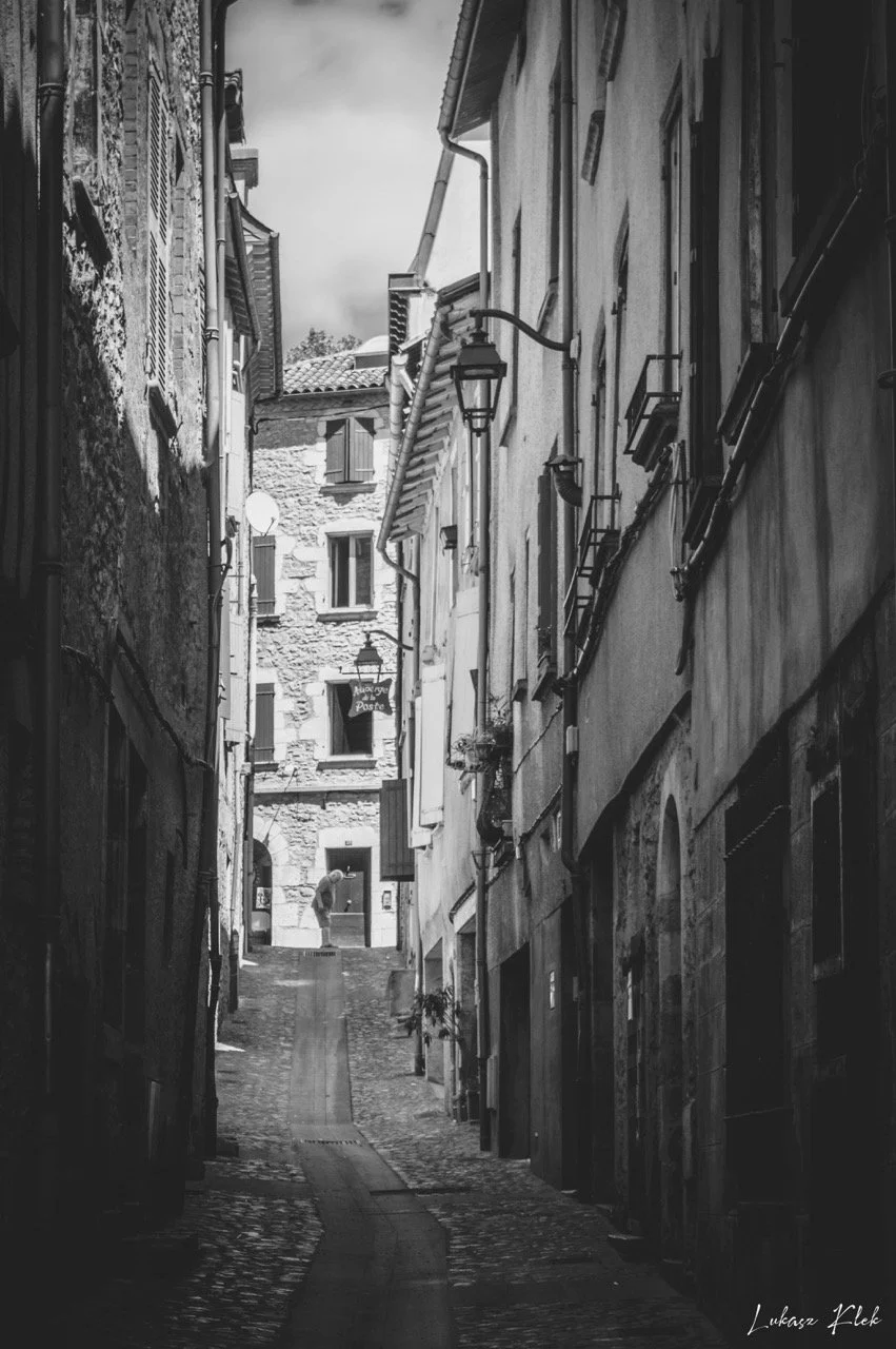 A narrow cobblestone alleyway in an old European town with tall buildings on both sides, featuring shuttered windows, street lamps, and a person walking in the distance.